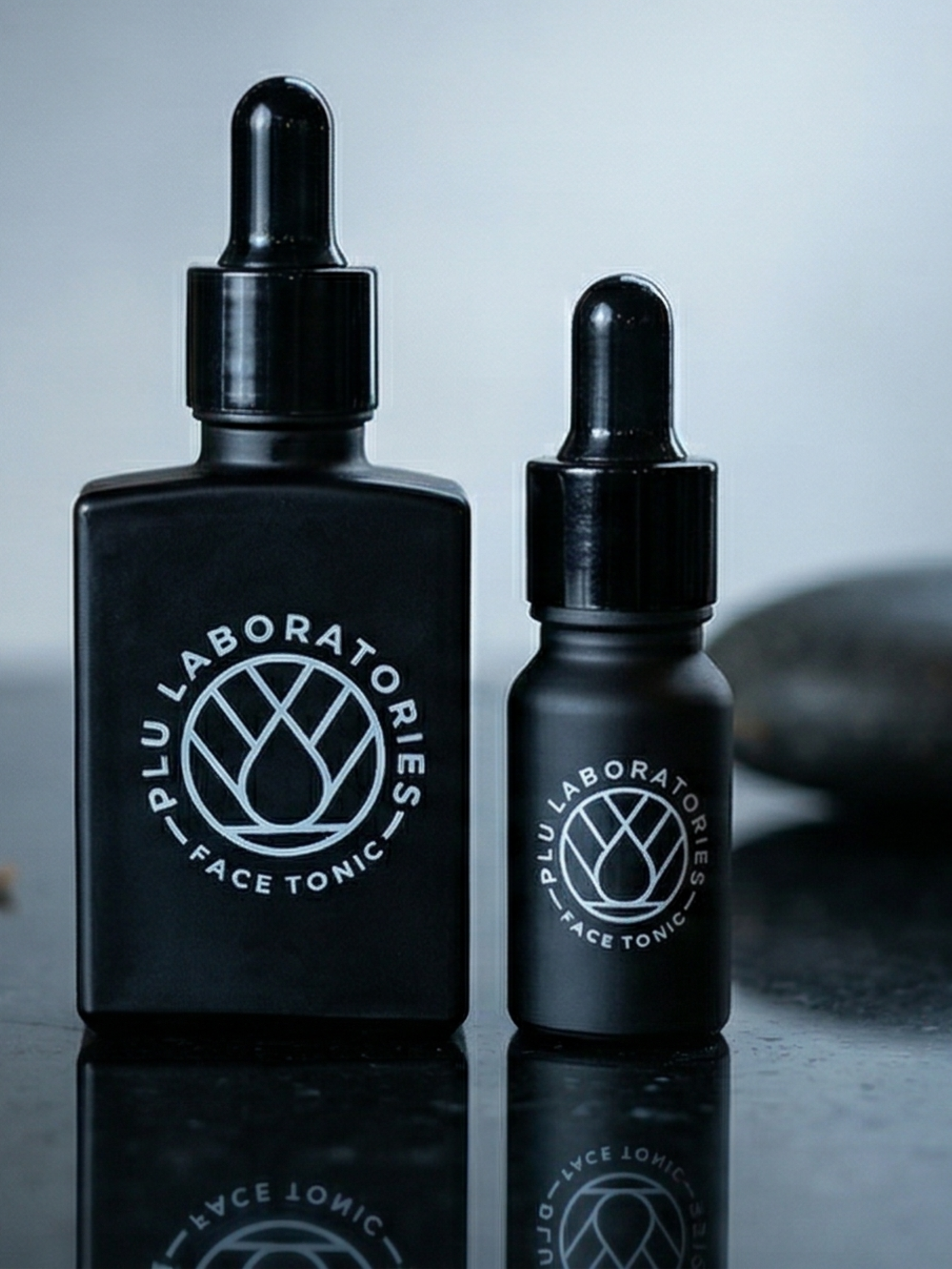 Two black bottles of Plu Laboratories Face Tonic on a reflective surface with a blurred background.